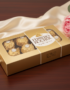 Caja dorada de bombones envueltos en papel oro sobre mesa de madera, junto a tela beige satinada y ramo de flores rosas y blancas