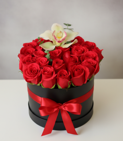 Arreglo de rosas rojas en caja redonda negra con lazo rojo y una orquídea blanca encima sobre fondo neutro