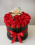 Arreglo de rosas rojas en caja redonda negra con lazo rojo y una orquídea blanca encima sobre fondo neutro