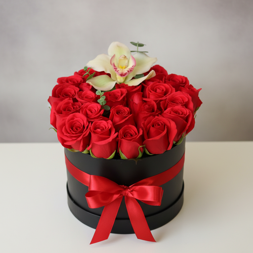 Arreglo de rosas rojas en caja redonda negra con lazo rojo y una orquídea blanca encima sobre fondo neutro
