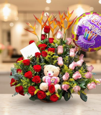 Arreglo de flores con rosas rojas y rosas, flores exóticas naranjas, peluche de oso y globo de feliz cumpleaños sobre mesa en interior