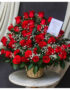 Canasta de mimbre sobre mesa redonda con gran arreglo de rosas rojas y flores blancas de relleno, acompañadas de tarjeta sobre fondo de cortinas blancas y tela gris