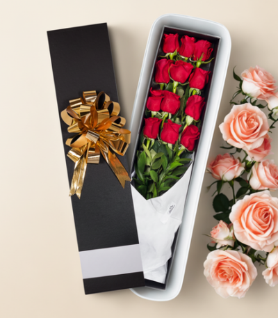 Caja de regalo alargada negra con lazo dorado que contiene un ramo de rosas rojas envueltas en papel blanco, junto a rosas rosadas sobre fondo beige