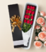 Caja de regalo alargada negra con lazo dorado que contiene un ramo de rosas rojas envueltas en papel blanco, junto a rosas rosadas sobre fondo beige