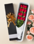 Caja de regalo alargada negra con lazo dorado que contiene un ramo de rosas rojas envueltas en papel blanco, junto a rosas rosadas sobre fondo beige