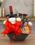 Arreglo floral AMADEA con rosas blancas, eucalipto, lavanda, vino tinto, chocolates variados y lazo rojo en cesta negra.