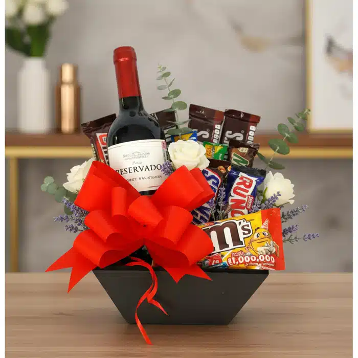 AMADEA: Arreglo Floral con Vino y Chocolates para Sorprender Arreglo floral AMADEA con rosas blancas, eucalipto, lavanda, vino tinto, chocolates variados y lazo rojo en cesta negra.
