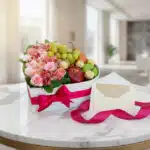 Arreglo floral AMOS en caja corazón con rosas, alstroemerias, frutas frescas y chocolates Ferrero Rocher, sobre mesa de mármo