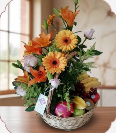 Cesta de mimbre con arreglo floral de rosas, lirios y gerberas, acompañada de frutas frescas como plátanos, uvas, manzanas y
