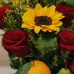 Primer plano extremo del Arreglo Floral Adriano con rosas rojas, girasol central, follaje verde y frutas frescas como naranja