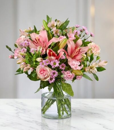 Arreglo floral ANNA con rosas y lirios rosados, alstroemerias y margaritas, en un jarrón de cristal sobre una mesa de mármol