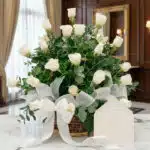 Arreglo floral blanco puro con rosas y follaje verde en una cesta de mimbre, decorado con una cinta de tela satinada blanca,