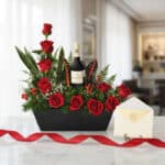Arreglo floral CINZIA con 12 rosas rojas, follaje verde, bayas rojas y una botella de vino tinto, presentado en una base negr