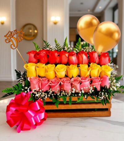 Arreglo floral Escala con rosas rojas, amarillas y rosadas en caja de madera, con globos dorados y lazo rosa, sobre mesa de m