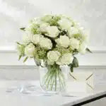 Arreglo floral Estefanía con 24 rosas blancas frescas en jarrón de cristal, tarjeta de regalo y cinta satinada sobre mármol.