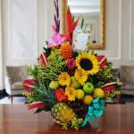 Arreglo floral exótico Antídoto con girasoles, heliconias, gladiolos, piña, uvas, peras y manzanas sobre mesa de madera pulid