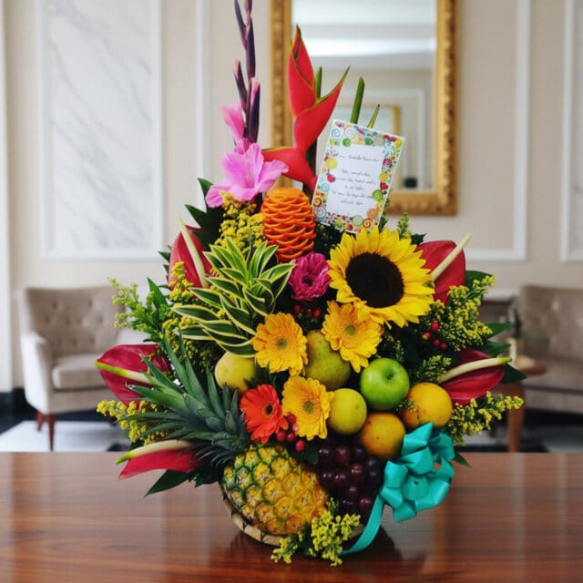 Arreglo floral exótico Antídoto con girasoles, heliconias, gladiolos, piña, uvas, peras y manzanas sobre mesa de madera pulid