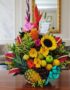 Arreglo floral exótico Antídoto con girasoles, heliconias, gladiolos, piña, uvas, peras y manzanas sobre mesa de madera pulid