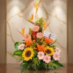 Arreglo floral exótico Columba con aves del paraíso, girasoles, lirios naranjas y margaritas rosadas sobre una mesa de madera
