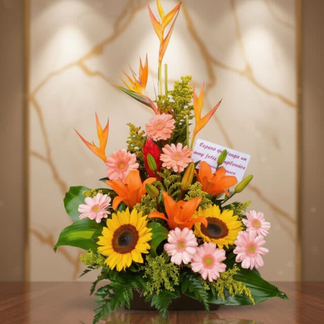 Arreglo floral exótico Columba con aves del paraíso, girasoles, lirios naranjas y margaritas rosadas sobre una mesa de madera
