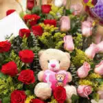 Primer plano extremo de Arreglo Floral Exótico Especial con rosas rojas y rosadas, follaje verde, gotas de rocío y un osito d