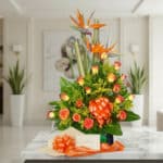 Arreglo floral exótico Esplendor con rosas naranjas y aves del paraíso, presentado en un entorno de lujo con una cinta decora