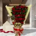 Arreglo floral de rosas rojas con chocolates Ferrero Rocher y tarjeta de regalo en un elegante entorno de mármol.