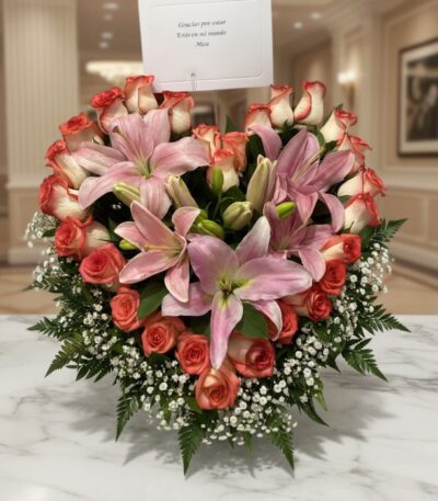 Arreglo floral Feliz Contigo con lirios rosados y rosas bicolor sobre mesa de mármol, fondo elegante de lobby de hotel boutiq