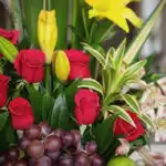 Primer plano extremo de Arreglo Floral con Frutas Akebia, destacando rosas rojas, lirios amarillos, uvas y hojas verdes con g