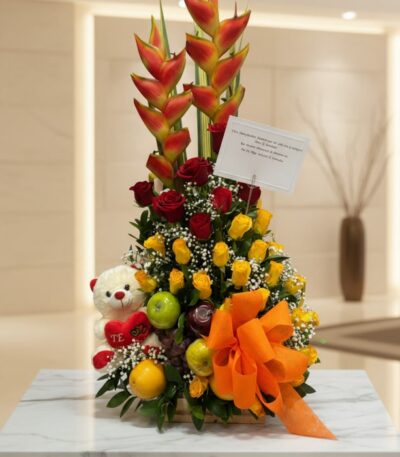 Arreglo floral premium con rosas rojas y amarillas, heliconias, frutas frescas y un lazo naranja, sobre una mesa de mármol en
