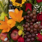 Primer plano extremo de un arreglo floral con frutas, destacando lirios naranjas, rosas rojas, uvas, manzanas y una botella d