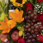 Primer plano extremo de un arreglo floral con frutas, destacando lirios naranjas, rosas rojas, uvas, manzanas y una botella d