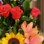 Primer plano extremo de un arreglo floral con rosas rojas, girasol, lirios naranjas y hojas verdes, destacando texturas y got