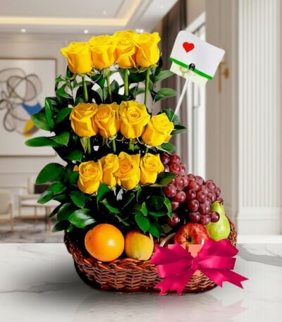 Arreglo floral con rosas amarillas, uvas rojas, manzana, pera y naranja en una cesta de mimbre con lazo rosa, sobre mármol.