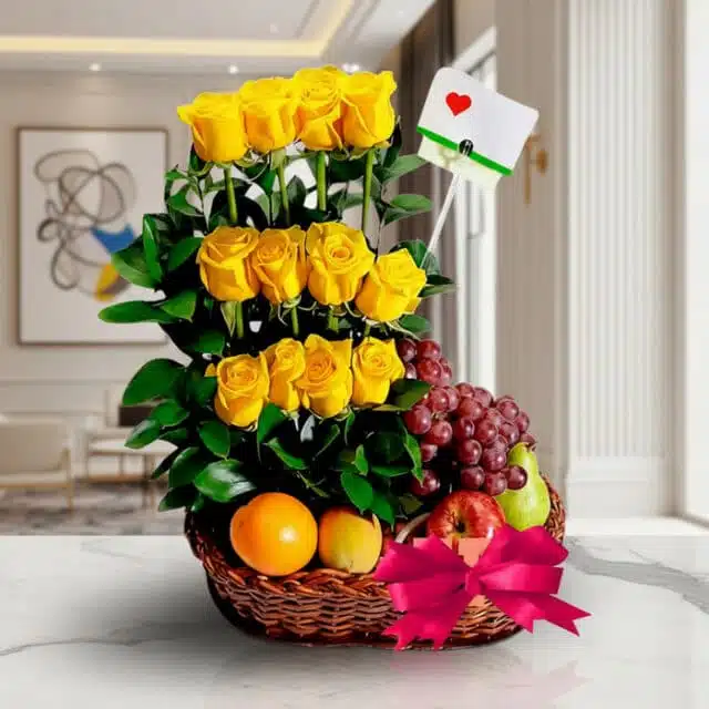 Arreglo floral con rosas amarillas, uvas rojas, manzana, pera y naranja en una cesta de mimbre con lazo rosa, sobre mármol.