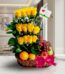 Arreglo floral con rosas amarillas, uvas rojas, manzana, pera y naranja en una cesta de mimbre con lazo rosa, sobre mármol.