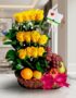 Arreglo floral con rosas amarillas, uvas rojas, manzana, pera y naranja en una cesta de mimbre con lazo rosa, sobre mármol.