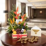 Arreglo floral exótico con flores de ave del paraíso y heliconias, piñas, uvas, manzanas y naranjas, acompañado de una botell
