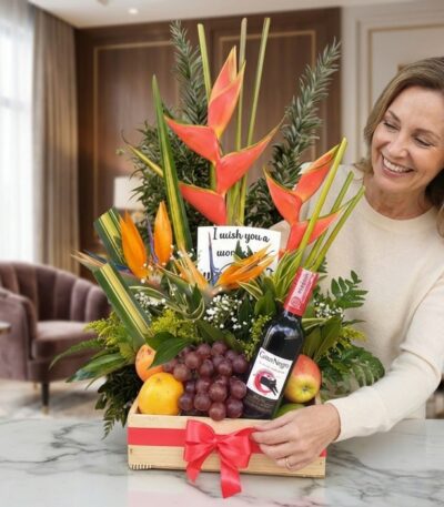 Mujer sonriente admirando un arreglo floral con heliconias, frutas frescas y una botella de vino tinto, presentado en una caj