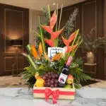 Arreglo floral Platonia con heliconias, aves del paraíso, uvas, manzanas, duraznos y botella de vino tinto, sobre mármol en u