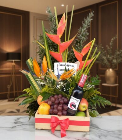 Arreglo floral Platonia con heliconias, aves del paraíso, uvas, manzanas, duraznos y botella de vino tinto, sobre mármol en u