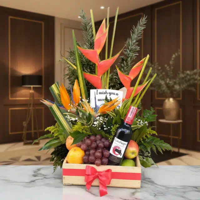 Arreglo floral Platonia con heliconias, aves del paraíso, uvas, manzanas, duraznos y botella de vino tinto, sobre mármol en u