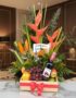 Arreglo floral Platonia con heliconias, aves del paraíso, uvas, manzanas, duraznos y botella de vino tinto, sobre mármol en u