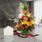Arreglo floral con rosas rojas, lirios amarillos, gerberas naranjas y frutas variadas en una cesta blanca, sobre una superfic