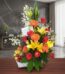 Arreglo floral con rosas rojas, lirios amarillos, gerberas naranjas y frutas frescas en una cesta blanca, sobre una mesa de m