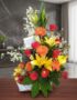 Arreglo floral con rosas rojas, lirios amarillos, gerberas naranjas y frutas frescas en una cesta blanca, sobre una mesa de m