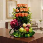 Arreglo floral premium con rosas salmón, uvas, manzanas, duraznos y peras, sobre una mesa de madera pulida en un lobby de hot