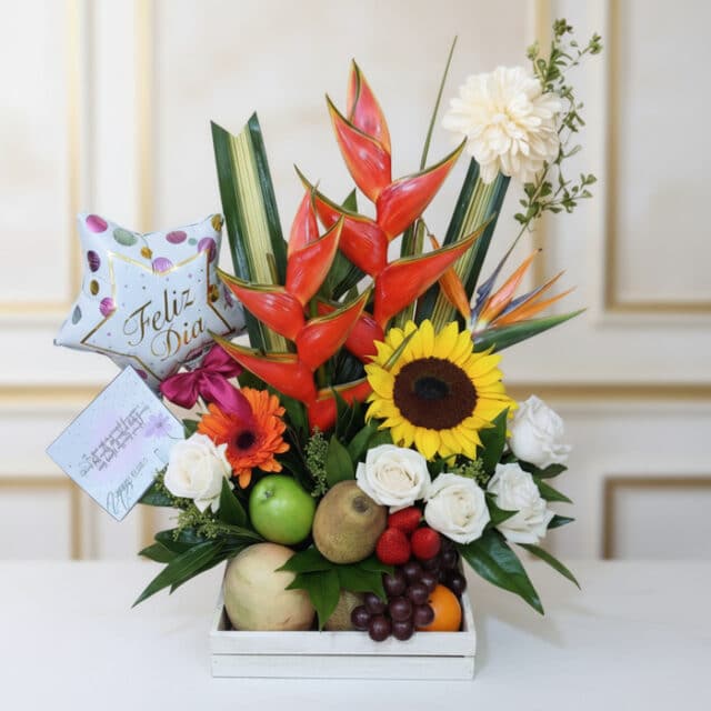 Arreglo floral con frutas tamarillo, girasoles, rosas blancas, heliconias y globo 'Feliz Día' en base de madera, ideal para e