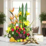 Arreglo floral Akebia con aves del paraíso, rosas rojas, lirios amarillos y frutas tropicales sobre mármol, listo para regalo