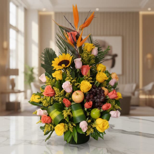 Arreglo floral vibrante con rosas amarillas y salmón, girasol, ave del paraíso y frutas tropicales como uvas, manzana y lima,